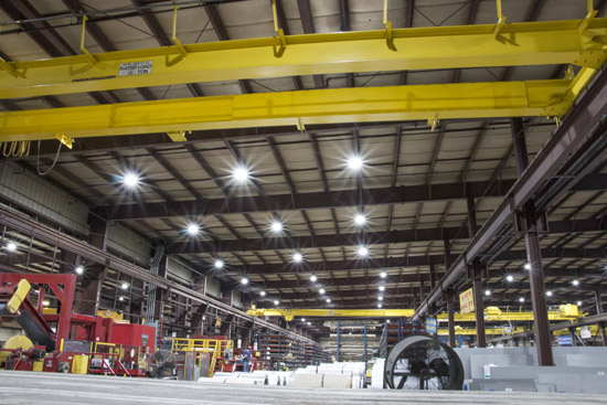 Overhead high bay LED lighting fixtures hanging down from a high ceiling warehouse with metal fabrication equipment and supplies, including large overhead cranes and I-beam support structures