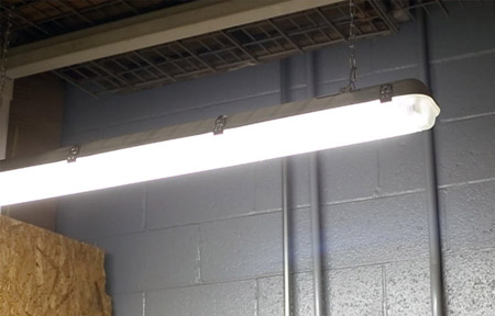 LED Vapor Tight Light Fixture hanging from overhead shelf illuminates a workspace with a grey cinder block wall