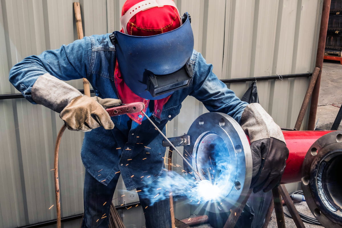 A pipefitter is shown using a stick welder to affix a mating flange on a piece of industrial pipe while the work area is illuminated by LED lights